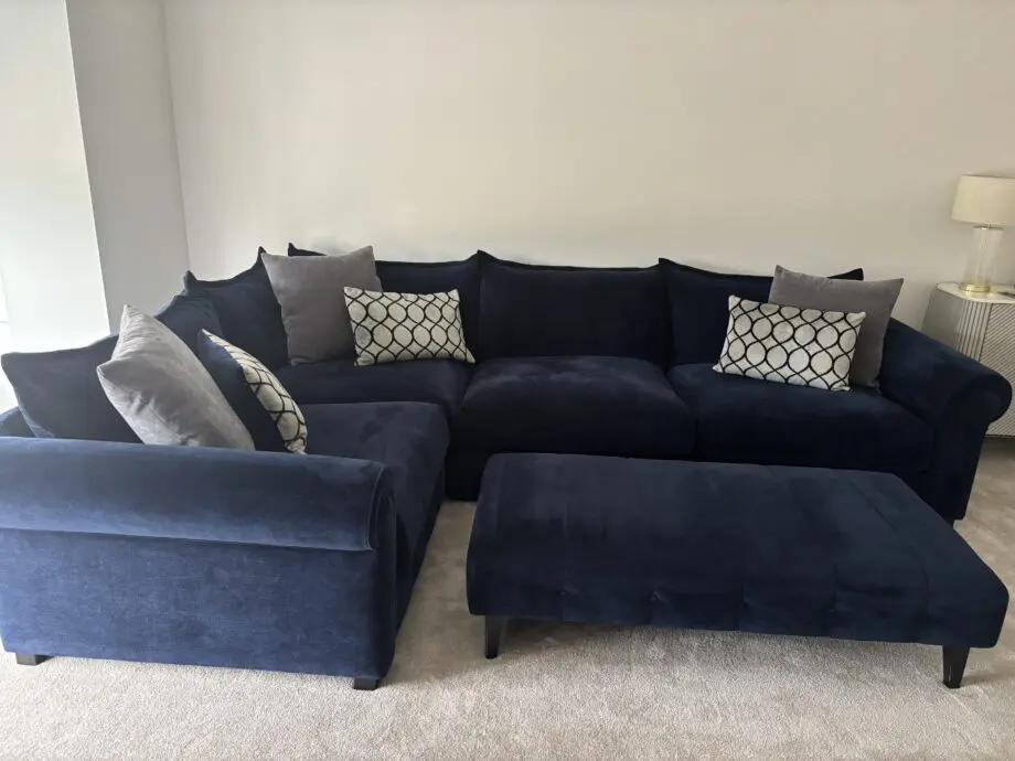 Large left hand corner sofa from Furniture Village. Navy blue with navy and grey scatter cushions included. Black feet. Matching footstool